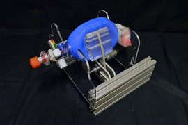 The new device fits around an Ambu bag (blue), which hospitals already have on hand in abundance. Designed to be squeezed by hand, instead they are squeezed by mechanical paddles (center) driven by a small motor. This directs air through a tube which is placed in the patient's airway.