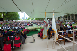 View of the many rows of people seated on Killian Court; the Commencement stage is in the foreground, with L. Rafael Reif speaking at the podium.