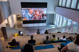 The amphitheater has 10 people sitting on the large wooden steps, and they watch Laura Dern and Sam Neill in “Jurassic Park” on a giant screen. Sunny windows and white walls surround the amphitheater.