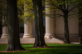 A student sits on the steps to an entrance to MIT. Large columns are behind her, and trees are in the foreground.