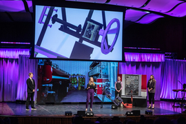 Members of the Purple Team wear purple and black on stage. Their self-spooling hose is on stage and a screen behind them shows their self-spooling hose and the inside of a firehouse.