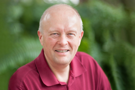 Peter Dedon portrait on blurry green background. Dedon is wearing a red shirt.