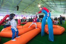 Under a large tent, a man on tall stilts wearing a clown-like outfit reaches down to high-five one of two young children climbing on an orange play structure.