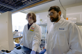 Two people in lab coats and blue gloves are in the lab, looking at a screen. On left, Miles wears a rainbow nametag that says “Miles,” and Goel’s coat says “Hansen Lab.”