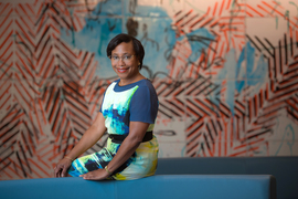 Paula Hammond poses for a portrait while sitting on the side of a couch. A mural with blue and orange is in blurry background.