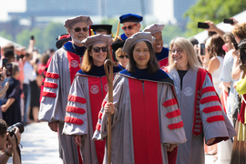 MIT faculty chair Lily Tsai, along with administration and faculty proceed through Killian Court. All are wearing academic regalia and Tsai holds a wooden staff.