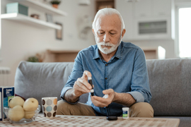 A man checking his blood-glucose levels on left index finger while sitting on gray couch.
