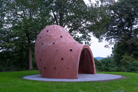 A side view of the red brick sculpture. It has an arched entryway, and then curves upward to an off-center dome ceiling. It has small circular windows scattered throughout.