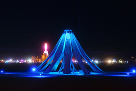 Night view of the Living Knitwork Pavilion, a pole with sheets of fabric draped over like a teepeee, that is lit in bright blue. Many points of light, from other parts of the Burning Man festival, appear in the background.