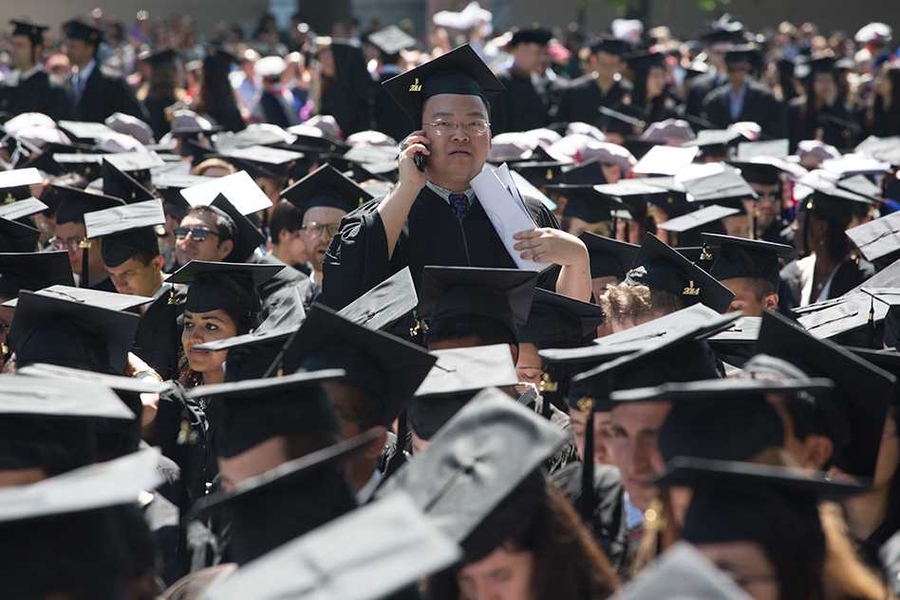 Graduating class at MIT commencement as one student stands up and is talking on their cell phone.