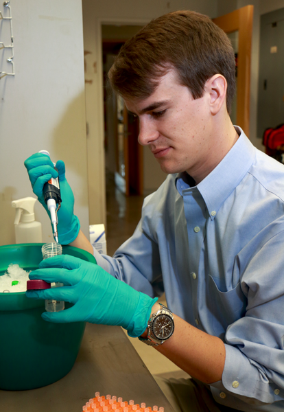 MIT Department of Physics graduate student James Owen Andrews works at a bench in the Cissé Lab, where he inserts genetic code for green fluorescent protein into living cells. The newly inserted code causes the target molecule to glow in a specific color that helps identify its location in fluorescent microscope images. Enzymes are chilled in an ice bucket (green container). Andrews is studying g...
