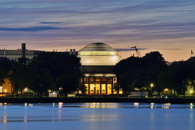 Photo of the MIT Great Dome from the vantagepoint of the Charles River, lit up with a sunset behind it and very still waters of the Charles in front of it