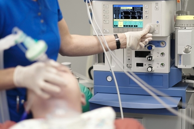 Photo of an anesthesiologist using the right hand to hold on a patient's breathing mask and the left hand to adjust settings on a monitor