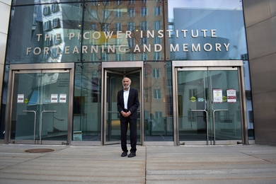 Photo of Emery Brown wearing a dark suit and white shirt standing in front of the glassy front entrance of the Picower Institute