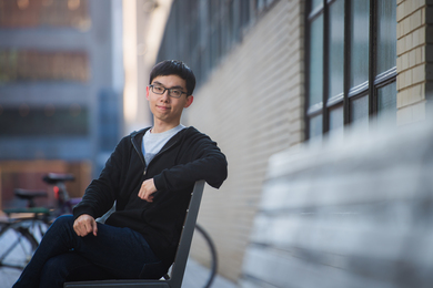 Changhao Li, photographed sitting outdoors on a bench next to a tan brick MIT building
