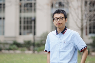 Hao Zhang poses for a photo in a grassy area in front of a stone MIT building