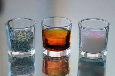 Three small glasses on a table hold (left to right), tiny silver-colored aluminum beads, a brownish orange liquid, and white salt crystals