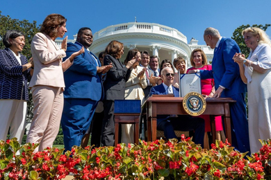 President Biden sits at a desk, while Senator Schumer stands next to the desk, propping up a signed document to face the camera. A row of people stands behind the president, standing and applauding, and the White House is in the background.