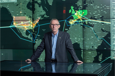 Photo of James Rice standing behind and leaning on a glass table. Behind him is a large world map with supply routes highlighted