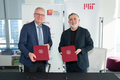 In a conference room, Christoph Meinel and Hashim Sarkis stand in front of banners with Hasso Plattner Institute and MIT logos. Each holds a red folder with the MIT seal on it.