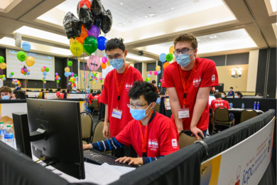 Sharing a single computer in a room filled with similar teams of coders, three students in matching orange T-shirts and face masks work on their coding solutions. 