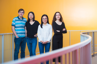 Photo of four students standing in a row on an interior curved walkway against an orange wall.