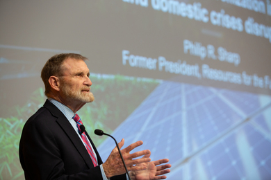 Philip R. Sharp speaking into a microphone in front of a projected slide showing out-of-focus solar cells