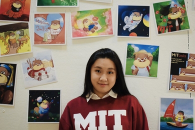 Photo of Margaret Zheng wearing an MIT sweatshirt with images of Wide Tim on the wall behind her