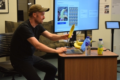 Andreas Refsgaard holds a banana and sits at a desk in front of computer screen showing images of the banana