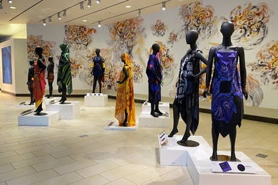 Nine mannequins on pedestals in a gallery, each wearing flowing designs in different brightly colored patterns