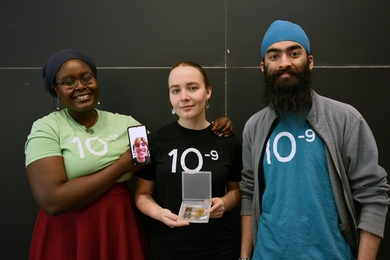 Three MIT students stand side by side wearing MIT.nano t-shirts that say "10^-9." The student in the center is holding a small plastic case featuring the electronic device they fabricated. The student on the left holds a smartphone with the face of another student from the group on the screen.