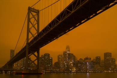 Photo of the Bay Bridge, from ground level, silhouetted against a dark orange sky with part of the San Francisco skyline in the background
