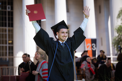 A male graduate wearing cap and gown, smiling and holding his diploma in the air