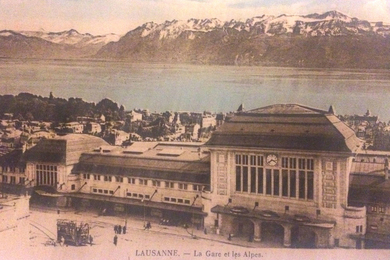 A pink-hued faded vintage postcard shows the buildings of Lausanne and the Alps in background, and says “LAUSANNE. La Gare et les Alpes.”