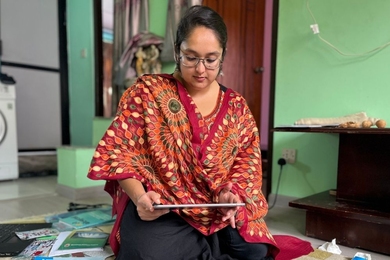 Apekshya Prasai kneels in a green room. Facing the camera, she looks down at the tablet computer she is holding.