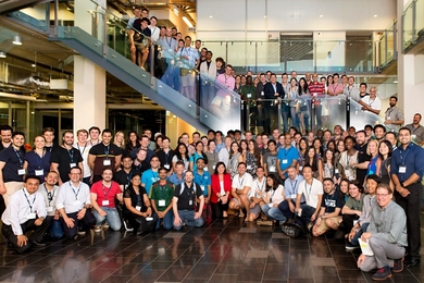 Hundreds of MIT Bootcamps alumni pose in a large atrium, near stairs. 