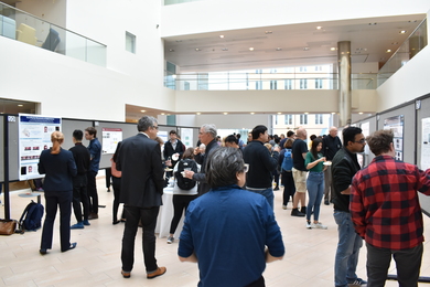 Scores of people mill about among tables of snacks and research posters on easels lining the walls in the large, brightly lit atrium of MIT Building 46 