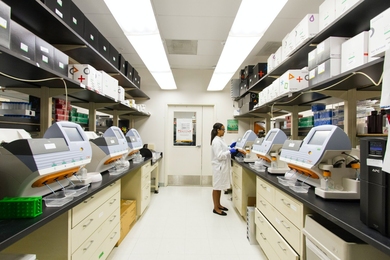 A woman in a white lab coat tends a bank of six machines in a lab