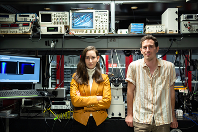 Madeleine Laitz, left, and Dane deQuilettes stand in a lab filled with equipment like computers, microscopes, and oscilloscopes.