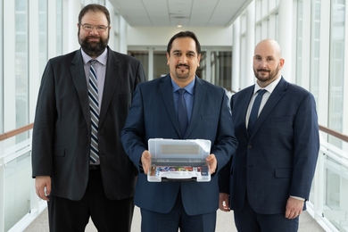David Bigelow, Hamed Okhravi, and Jason Martin stand together in a glass-lined hallway, with Okhravi holding a box containing a software package. 