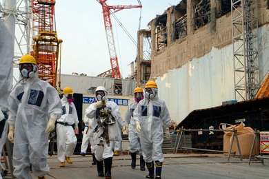 About 7 people wearing hard hats, white clean suits, and gas masks walk outside a nuclear power plant, with cranes and crumbling walls in the background.