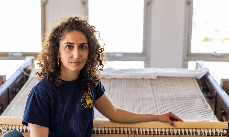 Chloe Bensahel sits in front of a loom, looking at the camera