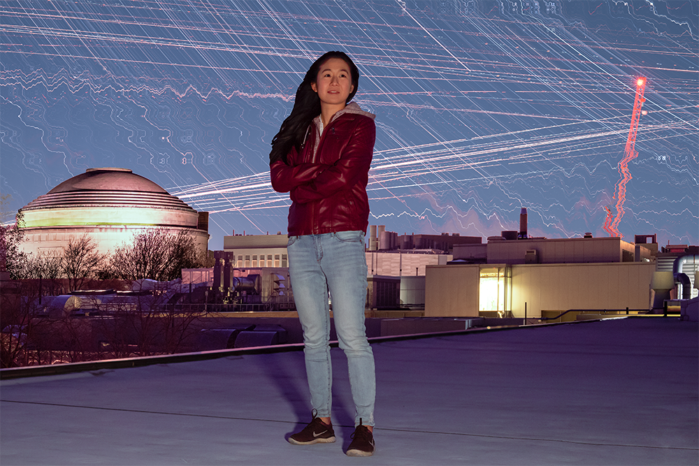 Juju Wang stands on a rooftop with airplane light trails in a bright night sky and the MIT dome behind her