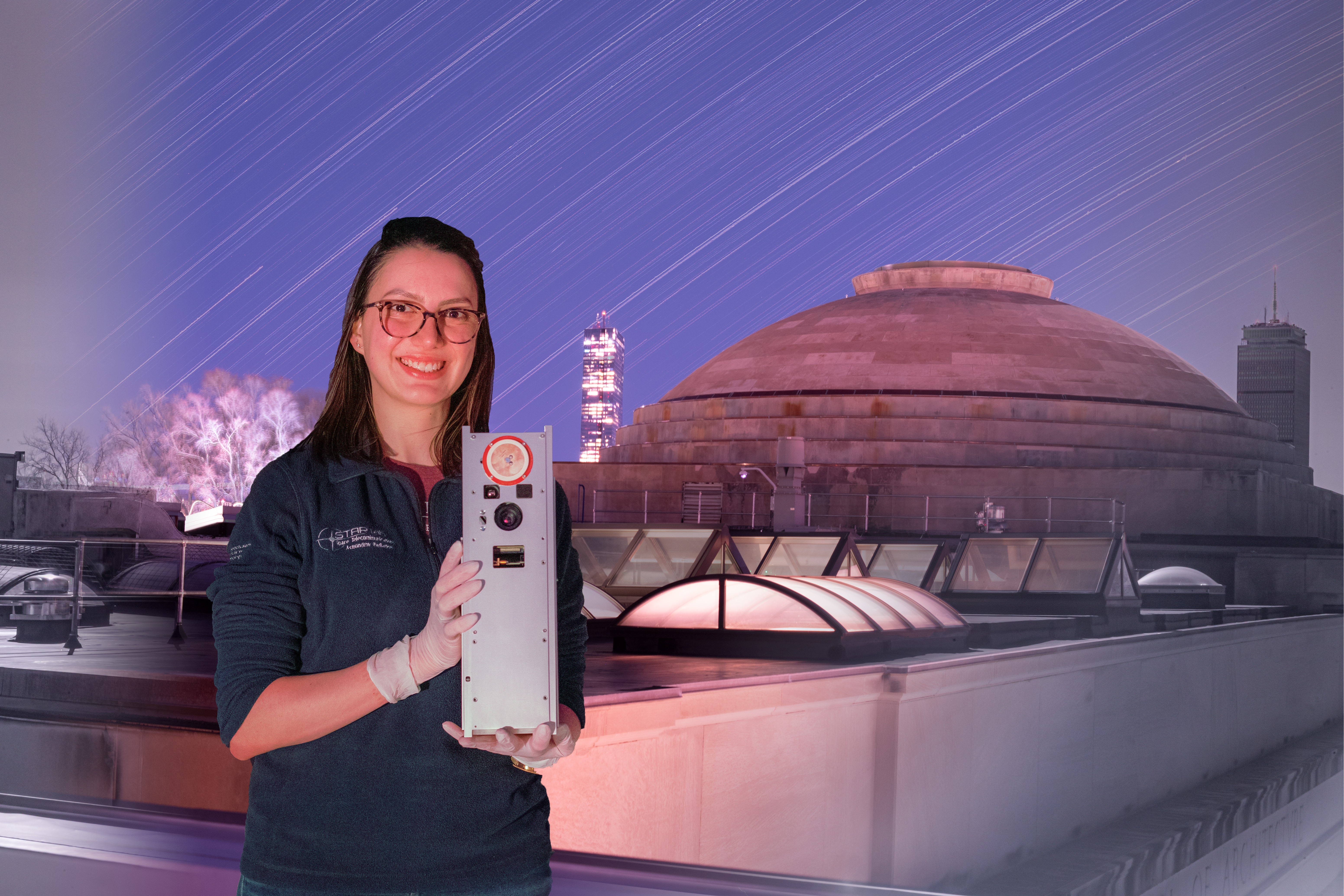 Paula do Vale Pereira, wearing latex gloves, holds BeaverCube, which is the size and shape of a desktop PC. Star trails are in the background, behind the MIT dome.
