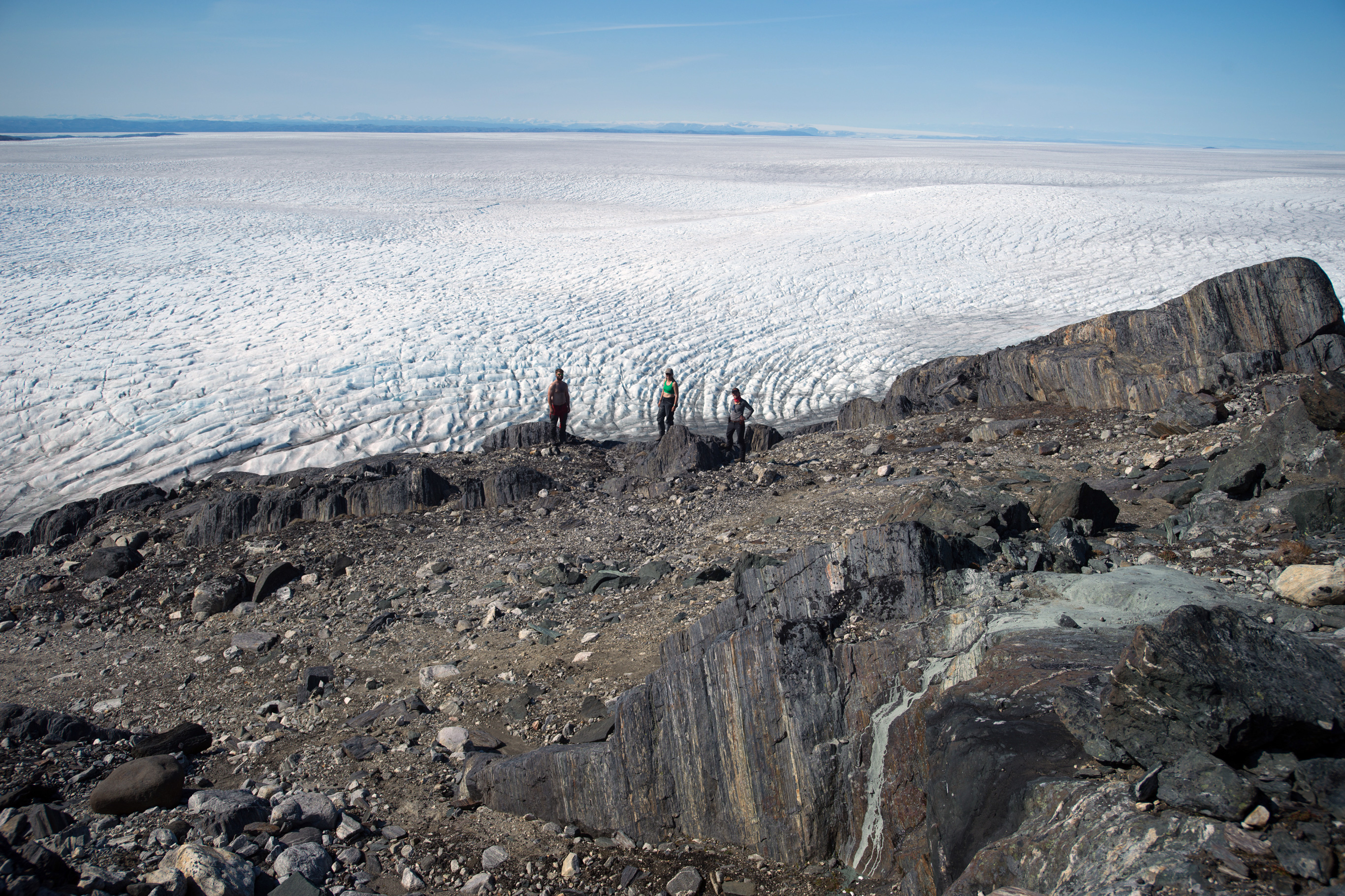 A drone photo shows three small researchers on a rocky formation, with a vast expanse of ice and snow in background.