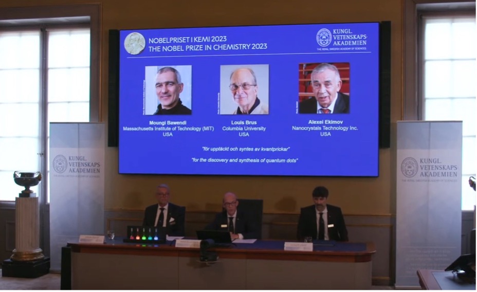 Three people seated at a table in conference room, with the Nobel Prize in Chemistry winners projected on screen behind them