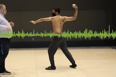 A man, left, watches as another man, shirtless and wearing electrodes on his skin, makes a pitching motion with his right arm cocked and his left arm extended toward his target