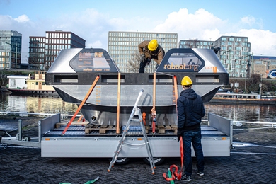 Photo of two men in hardhats working with a small metal boat on a trailer, next to a canal with buildings in the background