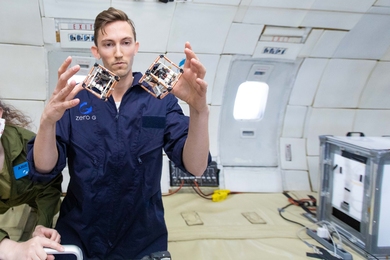 Photo of Martin Nisser in a blue jumpsuit inside a research airplane. Two cubes of electronics float between his hands.