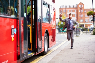 A man in a suit runs along a sidewalk with his arm raised, signaling to a red bus stopped along a curb.